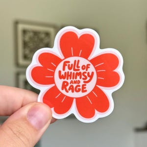 May include: A red flower-shaped sticker with a white border. The sticker has the words "Full of Whimsy and Rage" in a playful font. The sticker is held up by a hand.