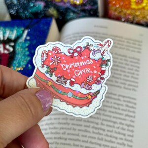 May include: A colorful Christmas-themed sticker in the shape of a cake. The sticker features a red cake with white frosting, snowflakes, a snowman, and the text "Christmas Girlie". Candy canes and presents are also included in the design.