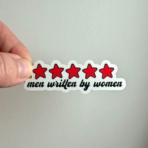May include: A white sticker with five red stars and the text "men written by women" in black script. The sticker is being held by a hand against a white background.