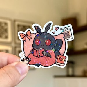 Peut inclure: Un autocollant holographique représentant un papillon de dessin animé lisant un livre. Le papillon est sur un oreiller et entouré de cœurs, d'un nœud, d'une tasse de café et d'un livre avec un cœur. Le texte "HAVE YOU SEEN ME?" est inclus.