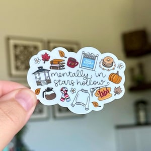 Peut inclure: Un autocollant blanc avec l'expression "mentally in stars hollow" et diverses illustrations, dont une cafetière, des livres, une tasse, un pull et une citrouille. L'autocollant a une forme de nuage et présente des images sur le thème de l'automne.
