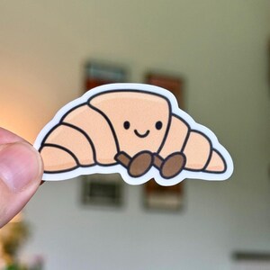 May include: A die-cut sticker featuring a light tan, cartoon-style croissant with a simple, smiling face. The croissant has dark brown, oval-shaped feet positioned at the bottom center, all outlined in a thick, clean white border.