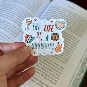 Könnte beinhalten: Ein holografischer Aufkleber mit dem Text "THE LIFE OF A SHOWGIRL" und Illustrationen eines Cocktails, eines Herzens, einer Discokugel und eines Badeanzugs. Der Aufkleber wird vor einem offenen Buch gehalten.