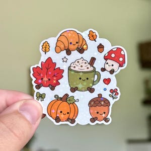 May include: A holographic sticker featuring cute, anthropomorphic autumn-themed illustrations including a croissant, a red maple leaf, a green mug of hot cocoa, a pumpkin, an acorn, and a red-and-white spotted mushroom, all with smiling faces.