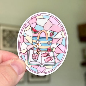 May include: A colorful sticker featuring a beach scene inside an oval stained-glass design. The image includes a striped tote bag, a book, a drink, flip-flops, and fruit. The color palette includes pink, blue, yellow, and white.