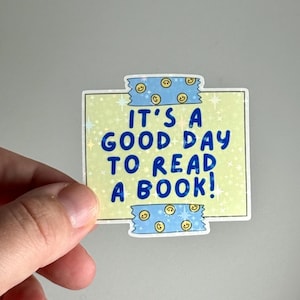 May include: A rectangular sticker with the text "IT'S A GOOD DAY TO READ A BOOK!" in blue. The sticker has a light yellow background with star accents and is held by a hand. Blue tape with smiley faces is at the top and bottom.