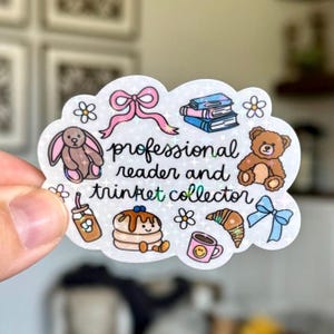May include: A holographic sticker in the shape of a cloud, featuring illustrations of a bunny, teddy bear, books, and food items. The sticker reads "professional reader and trinket collector" in black script.