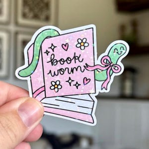 May include: A pink sticker featuring a book with the words "book worm" written on it. The book has a green worm with a pink bow wrapped around it. The sticker also has hearts, flowers, and stars.
