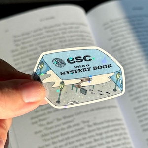 May include: A holographic sticker with the text "esc into a MYSTERY BOOK." The sticker features a fingerprint design, street lamps, a bench, and a briefcase. The sticker is held over an open book.