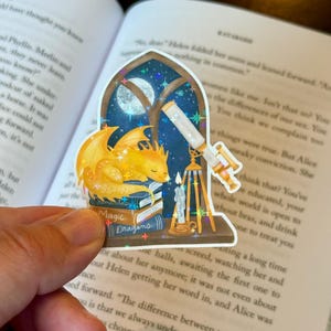 May include: A whimsical sticker featuring a sleeping yellow dragon atop a stack of books. The sticker includes a telescope, a candle, and the words "Fantasy Magic Dragons." The background shows a starry night sky.