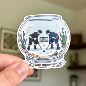 Pode incluir: Um autocolante com uma ilustração fantasiosa de dois jogadores de hóquei dentro de um aquário. Um jogador veste preto e dourado, o outro azul e vermelho. O texto "the boy aquarium" está abaixo.