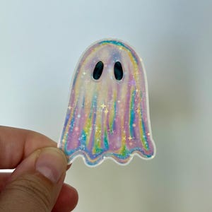 May include: A holographic ghost sticker with a pearlescent finish. The sticker features a spectral design with a rounded top and a wavy bottom edge. The ghost has two oval-shaped black eyes and is decorated with iridescent stars.