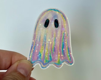 Op de afbeelding: Een holografische spooksticker met een parelmoer afwerking. De sticker heeft een spectraal ontwerp met een afgeronde bovenkant en een golvende onderrand. De geest heeft twee ovale zwarte ogen en is versierd met iriserende sterren.