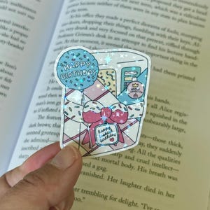 Puede incluir: Una pegatina holográfica con un diseño de caja de regalo de cumpleaños, un lazo rosa y chispas. La pegatina incluye el texto "Happy Birthday" en un globo azul y una etiqueta de regalo.