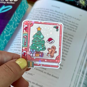 May include: A festive Christmas sticker with a book-shaped design. The sticker shows a Christmas tree, presents, a gingerbread man, and a Santa hat. The sticker has a holographic, glittery finish, adding a touch of sparkle to the design.