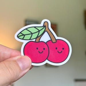 May include: A die-cut sticker featuring two bright red cherries with brown stems and a single green leaf. Each cherry has a simple black line drawing of a smiling face with two small dots for eyes and a curved line for a mouth, outlined in white.
