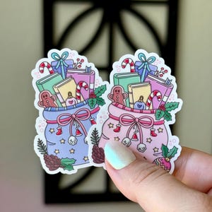 May include: Two stickers featuring bags filled with Christmas gifts. One bag is light blue, the other is pink. Each bag contains books, candy canes, gingerbread men, and ornaments. The bags are decorated with bows, stars, and pinecones.