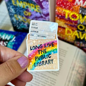 May include: A sticker with the text "LONG LIVE THE PUBLIC LIBRARY" in black, rainbow, and white. The sticker is in the shape of a library card and has a small smiley face and other small graphics. The sticker is held in front of an open book.