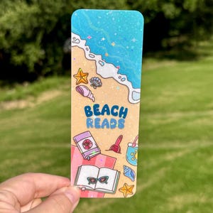 May include: A rectangular bookmark with a holographic finish featuring a beach scene with sand, blue ocean waves, seashells, and starfish. The text BEACH READS is printed in blue. Illustrations include a pink towel, a book, and sunglasses.
