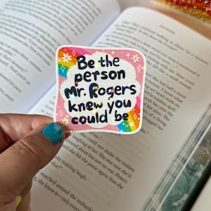 Op de afbeelding: Een vierkante sticker met de tekst "Be the person Mr. Rogers knew you could be" in het zwart, met een roze wolkenachtergrond en regenboogaccenten. De sticker wordt boven een open boek gehouden.