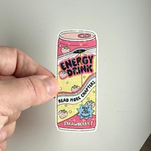 May include: A sticker in the shape of a pink and yellow energy drink can. The can features the words "ENERGY DRINK" and "STRAWBERRY" with strawberry illustrations. The text "READ MORE CHAPTERS" and "READ ALL NIGHT GUARANTEED" are also included.