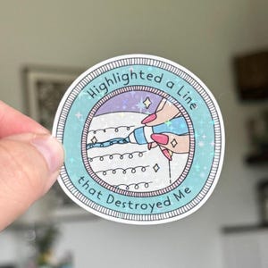 May include: A round sticker with the text "Highlighted a Line that Destroyed Me." The sticker features a hand with pink nails highlighting a line with a blue highlighter. The design has a light blue and purple color scheme.