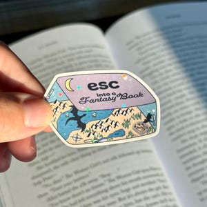 May include: A sticker with the text "esc into a Fantasy Book" featuring a fantasy map design with mountains, water, and a crescent moon. The sticker has a holographic finish and is held over an open book.