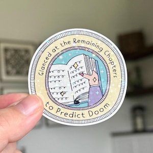 May include: A round sticker with the text "Glanced at the Remaining Chapters to Predict Doom." The sticker features an illustration of a hand holding an open book with stars and sparkles. The design has a light blue and yellow color scheme.