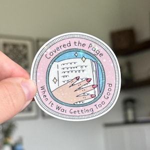 May include: A round sticker with the text "Covered the Page When It Was Getting Too Good." The sticker features a hand with pink nails covering an open book. The design is set within a pink and blue border with star accents.