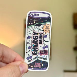 May include: A holographic sticker shaped like an energy drink can with a dark purple and white design. It features text reading ENERGY DRINK, READ ALL NIGHT, HORROR READER, READ MORE CHAPTERS, and DARK CHERRY, along with cherry and star graphics.