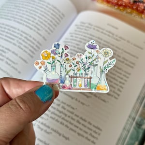 Puede incluir: Una colorida pegatina con un diseño floral de laboratorio. La pegatina muestra varias flores en vasos de precipitados y tubos de ensayo, con un arcoíris de colores. La pegatina se sostiene en una mano, con un libro en el fondo.