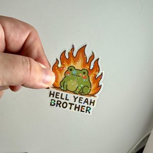 May include: A sticker depicting a green cartoon frog surrounded by flames, with the words "HELL YEAH BROTHER". The frog has sparkly details, and the flames are orange and yellow. The text is in a holographic font.