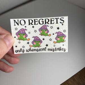 May include: A white sticker with a glittery finish featuring the text "NO REGRETS only whimsical mistakes". The sticker has a design of four green frogs wearing purple wizard hats and riding broomsticks. The frogs are surrounded by black stars.