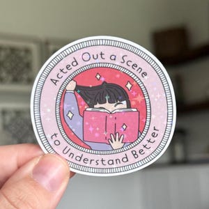 May include: A round sticker with a pink border and the text "Acted Out a Scene to Understand Better." The sticker features an illustration of a person reading a pink book, with stars and sparkles. The person is pulling their hair.