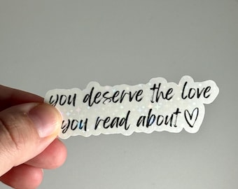 May include: A holographic sticker with the text "you deserve the love you read about" in black script. The sticker has a white background and a heart symbol at the end of the phrase.