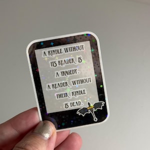 May include: A black and white sticker with a glittery background and a dragon design. The sticker says "A Kindle without its reader is a tragedy. A reader without their Kindle is dead."
