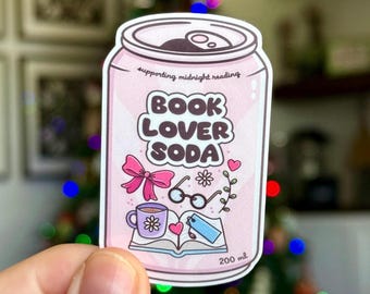 May include: A pink sticker in the shape of a soda can with the words "BOOK LOVER SODA." The sticker features illustrations of a bow, glasses, a cup of coffee, an open book, and a phone. The text "supporting midnight reading" is also included. The sticker is 200 ml.