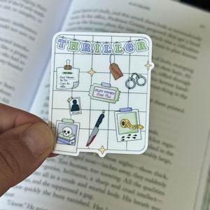 May include: A white sticker with the word "THRILLER" in a bubble font. The sticker has a grid design with images of a knife, handcuffs, and a skull. The sticker is held over an open book.
