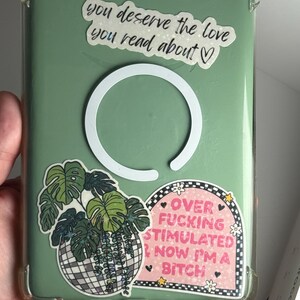 May include: A clear phone case with a green backing, decorated with stickers. One sticker reads "you deserve the love you read about." Another says "OVER FUCKING STIMULATED NOW I'M A BITCH." A third sticker features a plant in a disco ball.