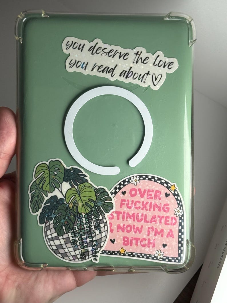 May include: A clear phone case with a green backing, decorated with stickers. One sticker reads "you deserve the love you read about." Another says "OVER FUCKING STIMULATED NOW I'M A BITCH." A third sticker features a plant in a disco ball.