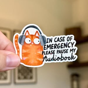 Op de afbeelding: Sticker met een oranje kat met een koptelefoon en de tekst "IN CASE OF EMERGENCY PLEASE PAUSE MY Audiobook". De sticker heeft een glitterafwerking.