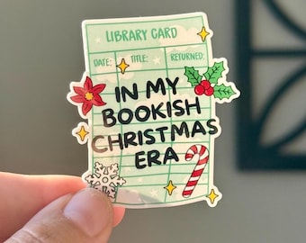 May include: A sticker in the shape of a library card with the text "IN MY BOOKISH CHRISTMAS ERA." The sticker has a light green background with Christmas-themed decorations, including a candy cane, holly, and a snowflake.