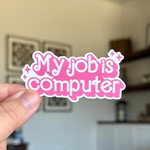 May include: A pink sticker with the words "My job is computer" in a whimsical font. The sticker has a white border and starburst details. The sticker is held up by a hand.