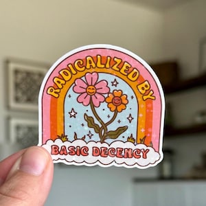 Op de afbeelding: Een kleurrijke sticker met een regenboogboog en de tekst "RADICALIZED BY BASIC DECENCY". De sticker toont twee lachende bloemen tegen een blauwe achtergrond met sterren.