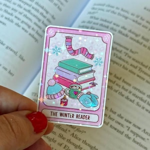 May include: A rectangular sticker with a pink border features a winter theme. It shows a stack of books, a scarf, a hat, mittens, a mug, a candle, and snowflakes. The text "THE WINTER READER" is at the bottom.