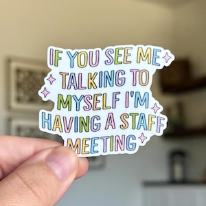 以下が含まれることがあります： カラフルな文字で「IF YOU SEE ME TALKING TO MYSELF I'M HAVING A STAFF MEETING」と書かれた白いステッカー。ステッカーはユニークな形で、手で持たれています。