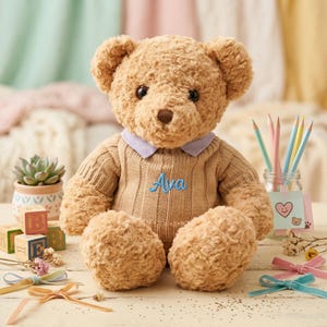 Può includere: Un orsacchiotto di peluche con un maglione marrone chiaro con il nome "Ava" ricamato in blu. La scena include blocchi di legno, matite e nastri, creando un'atmosfera calda e invitante.