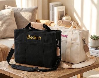 May include: Two rectangular canvas tote bags with adjustable shoulder straps and top handles featuring light tan leather accents. The black bag displays the name Beckett in yellow embroidery, while the cream bag shows a name in pink text.