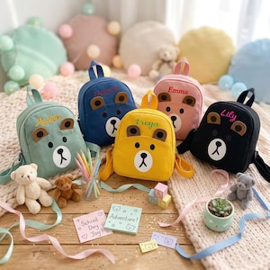 May include: A selection of bear-faced backpacks in shades of green, blue, yellow, pink, and black, each personalised with a name. The image also features teddy bears, pencils, and small decorative notes, creating a playful scene.