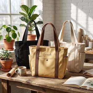 May include: Three canvas tote bags in black, beige, and cream, with side pockets and long handles. The beige bag has brown straps and the name "Sarah" embroidered on it. The bags are on a wooden table with plants and a cup.